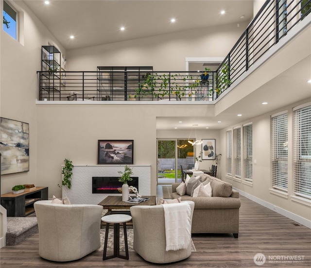 living room with a towering ceiling, a glass covered fireplace, a healthy amount of sunlight, wood finished floors, and baseboards