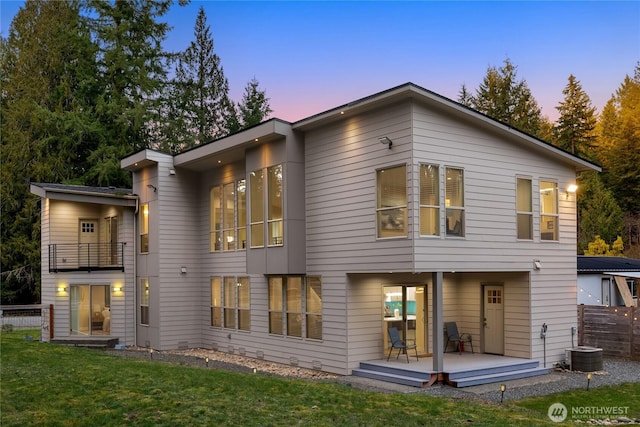 back of house at dusk featuring central AC unit, a balcony, fence, a lawn, and a wooden deck