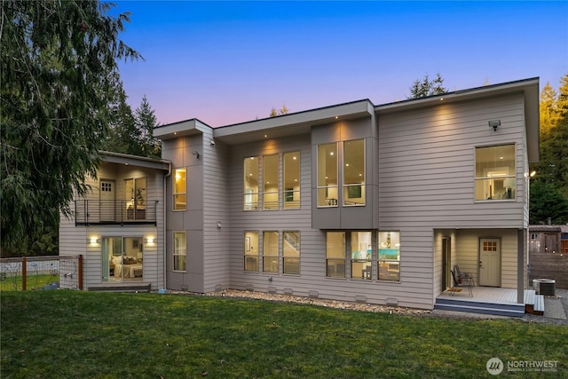 back of house at dusk featuring a balcony, fence, and a lawn