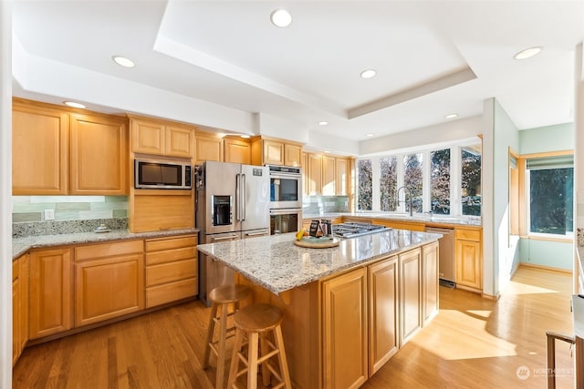 kitchen featuring stainless steel appliances, a center island, a tray ceiling, light hardwood / wood-style floors, and light stone countertops