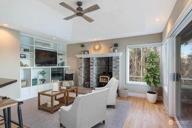 living room featuring a tray ceiling, a wood stove, ceiling fan, wood finished floors, and baseboards