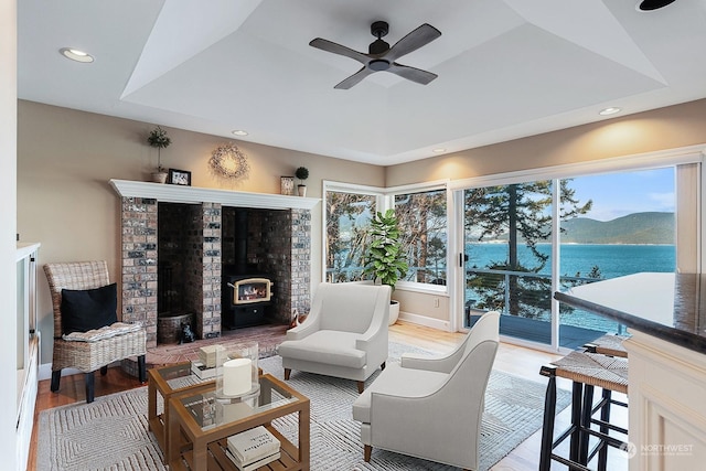 living area with a raised ceiling, a ceiling fan, a wood stove, light wood-style floors, and recessed lighting