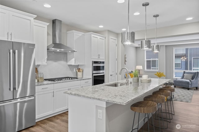 kitchen with sink, stainless steel appliances, an island with sink, white cabinets, and wall chimney exhaust hood