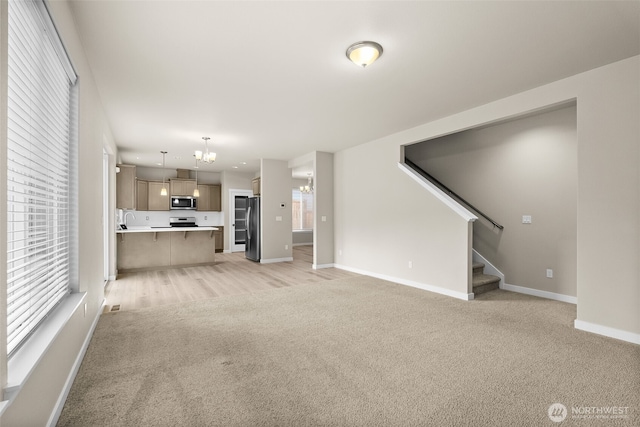 unfurnished living room with baseboards, an inviting chandelier, a sink, stairs, and light colored carpet