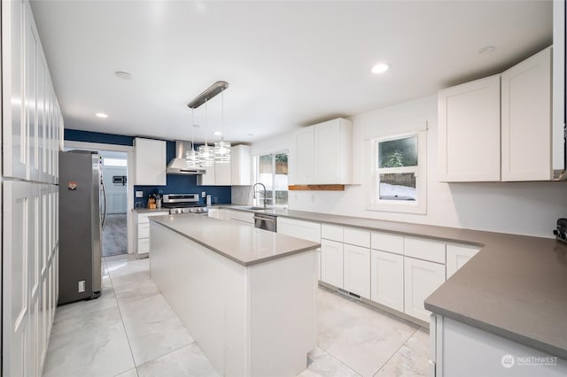 kitchen with wall chimney exhaust hood, white cabinetry, decorative light fixtures, appliances with stainless steel finishes, and a kitchen island