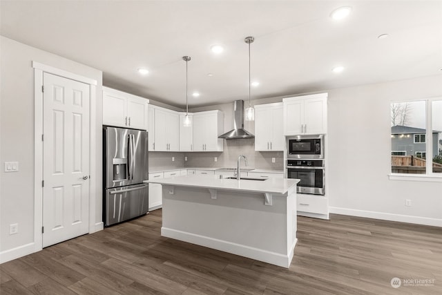 kitchen with appliances with stainless steel finishes, pendant lighting, white cabinetry, an island with sink, and wall chimney range hood