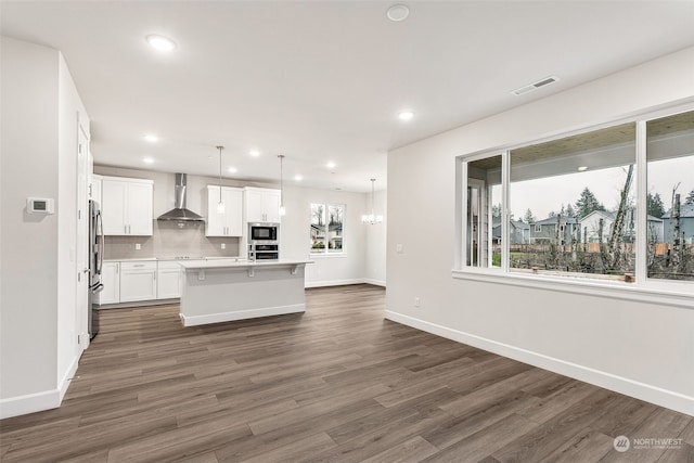 kitchen with wall chimney range hood, appliances with stainless steel finishes, white cabinetry, hanging light fixtures, and a center island with sink