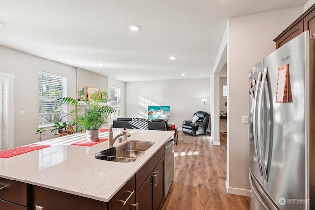 kitchen featuring sink, appliances with stainless steel finishes, a kitchen island with sink, dark brown cabinets, and light stone countertops