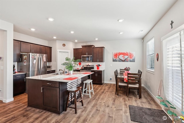 kitchen featuring appliances with stainless steel finishes, an island with sink, a breakfast bar area, dark brown cabinetry, and light wood-type flooring