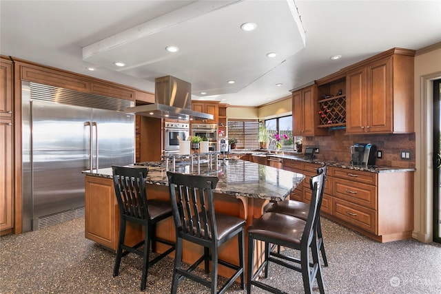 kitchen featuring appliances with stainless steel finishes, a breakfast bar, dark stone countertops, a large island with sink, and island exhaust hood