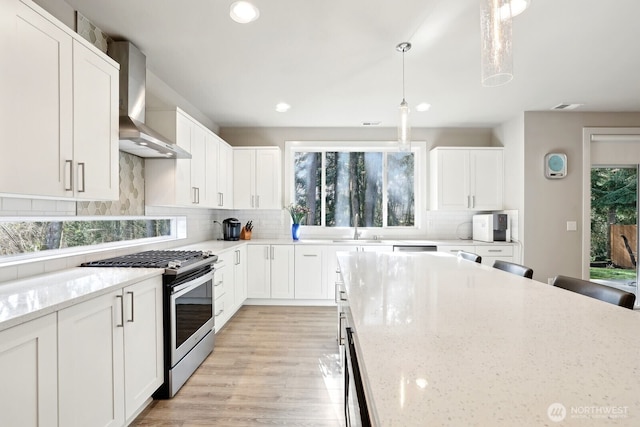 kitchen with tasteful backsplash, stainless steel gas range oven, white cabinets, wall chimney exhaust hood, and light stone counters