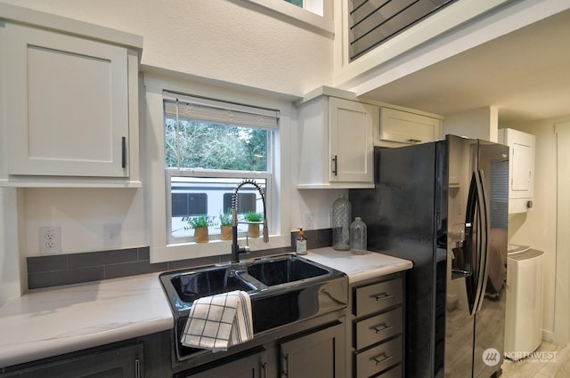 kitchen with sink, white cabinets, stacked washer / drying machine, light stone countertops, and black refrigerator with ice dispenser