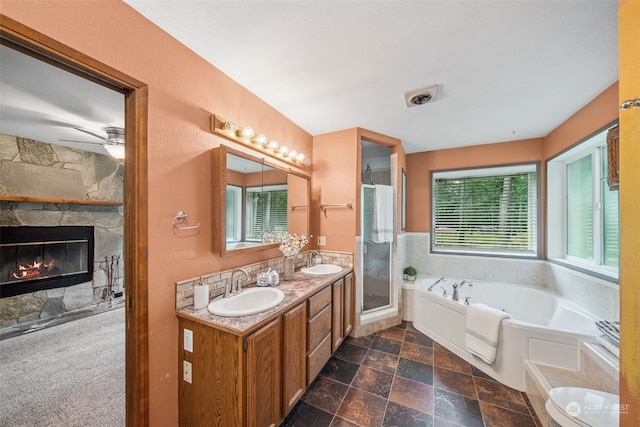 full bathroom featuring independent shower and bath, a large fireplace, vanity, ceiling fan, and toilet