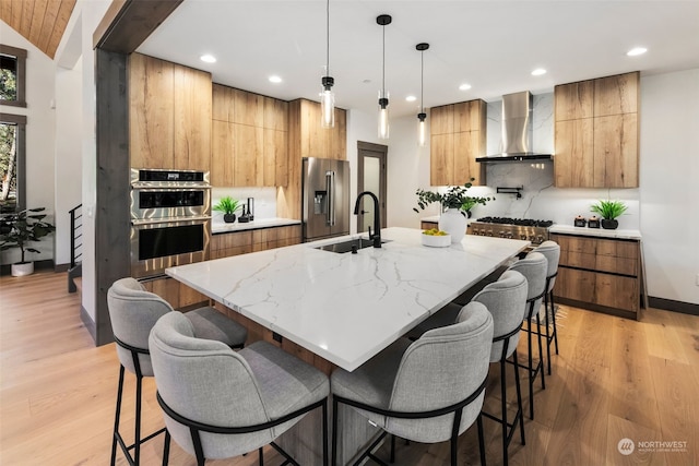 kitchen with pendant lighting, high quality appliances, an island with sink, sink, and wall chimney range hood