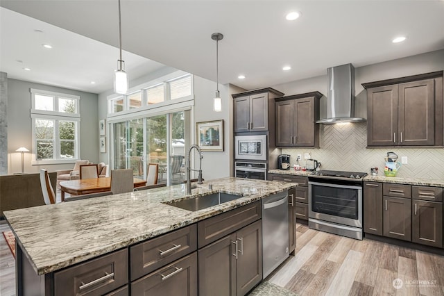 kitchen with appliances with stainless steel finishes, sink, light hardwood / wood-style flooring, wall chimney exhaust hood, and hanging light fixtures