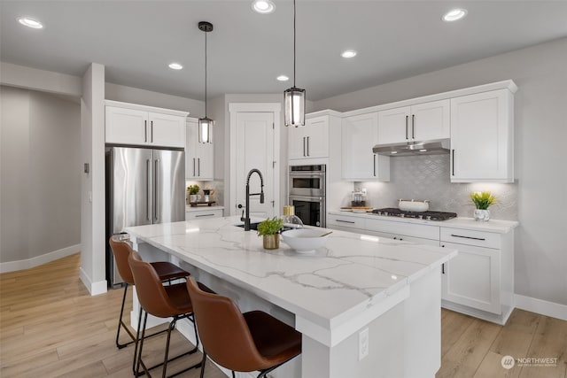 kitchen featuring white cabinetry, a center island with sink, pendant lighting, stainless steel appliances, and light stone countertops