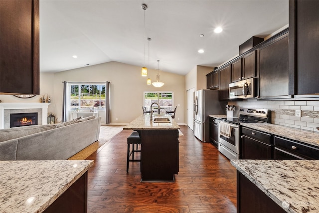 kitchen with lofted ceiling, appliances with stainless steel finishes, a kitchen breakfast bar, an island with sink, and light stone countertops