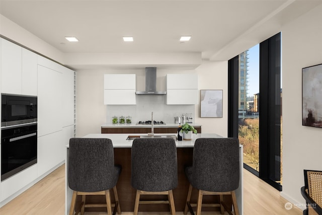 kitchen featuring stainless steel microwave, an island with sink, white cabinets, oven, and wall chimney exhaust hood