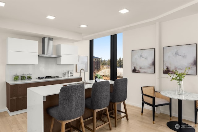 kitchen featuring wall chimney range hood, sink, a kitchen island with sink, gas stovetop, and white cabinets