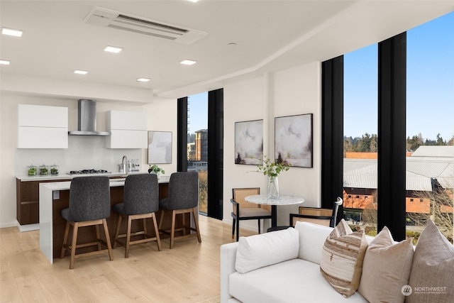 kitchen with white cabinets, wall chimney range hood, floor to ceiling windows, a center island with sink, and light wood-type flooring