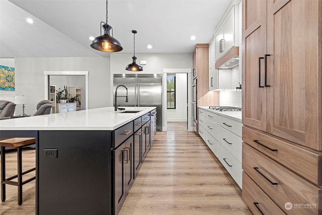 kitchen with appliances with stainless steel finishes, white cabinetry, a kitchen island with sink, decorative light fixtures, and light wood-type flooring