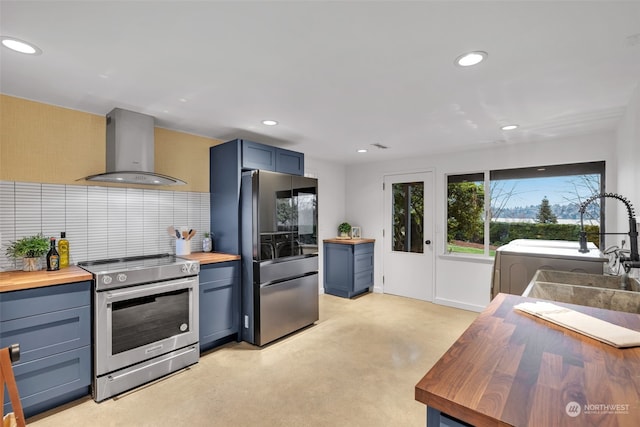 kitchen with appliances with stainless steel finishes, blue cabinets, wooden counters, backsplash, and wall chimney exhaust hood