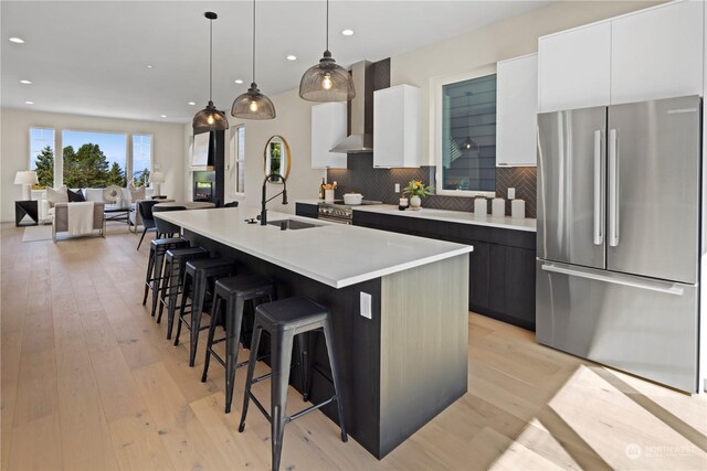 kitchen featuring appliances with stainless steel finishes, white cabinets, a center island with sink, decorative light fixtures, and wall chimney exhaust hood