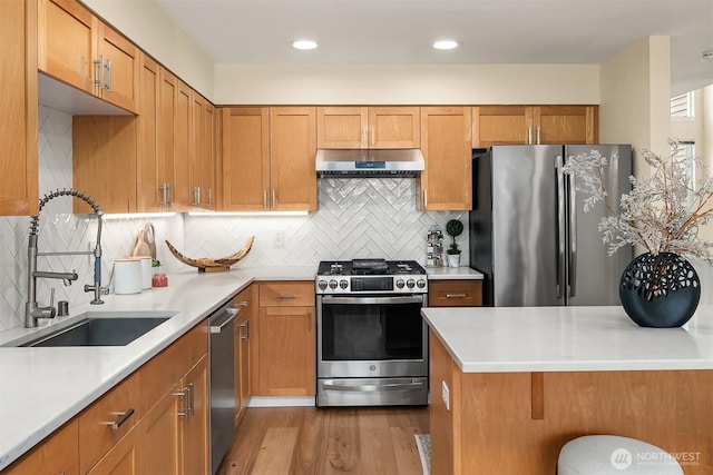 kitchen with brown cabinetry, a sink, stainless steel appliances, light wood-style floors, and under cabinet range hood