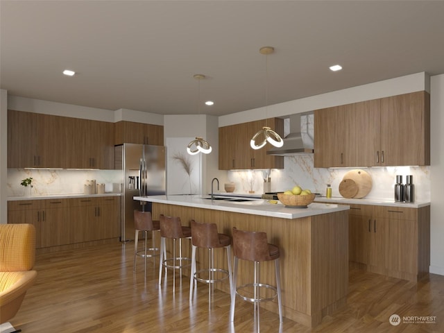 kitchen featuring a center island with sink, stainless steel fridge with ice dispenser, a breakfast bar area, light countertops, and wall chimney range hood