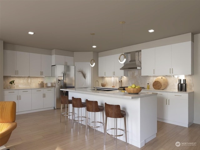 kitchen with wall chimney range hood, hanging light fixtures, white cabinets, and a center island with sink