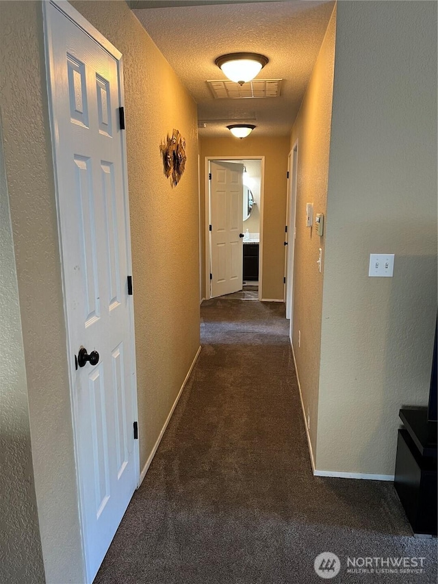 hallway featuring a textured ceiling, a textured wall, visible vents, baseboards, and dark colored carpet