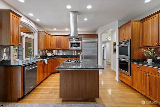 kitchen with dark stone counters, island exhaust hood, a center island, built in appliances, and light wood-type flooring