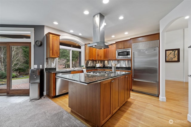 kitchen with a center island, built in appliances, island range hood, decorative backsplash, and dark stone counters