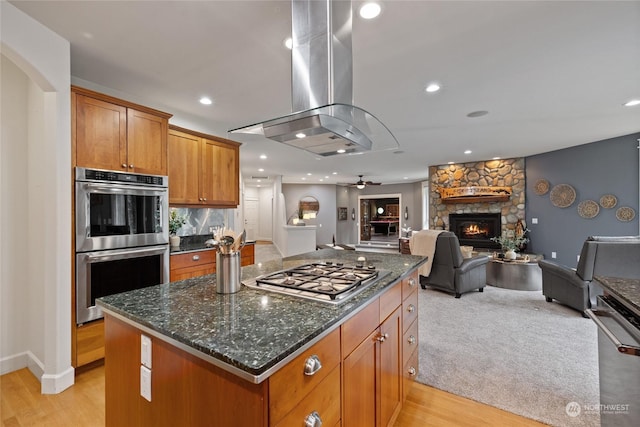 kitchen with appliances with stainless steel finishes, a stone fireplace, dark stone counters, island exhaust hood, and a center island