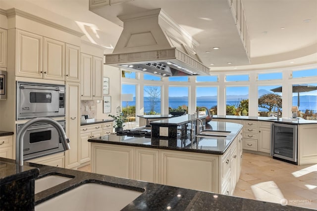 kitchen featuring a water view, dark stone countertops, island exhaust hood, beverage cooler, and a kitchen island with sink