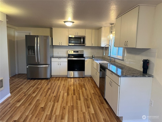 kitchen with appliances with stainless steel finishes, white cabinetry, dark stone counters, hanging light fixtures, and light hardwood / wood-style floors