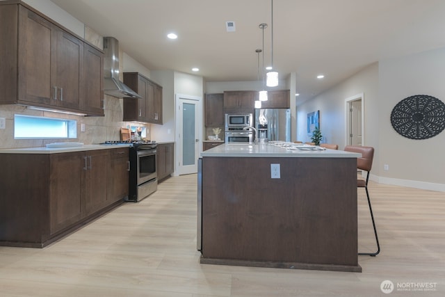 kitchen featuring dark brown cabinets, wall chimney range hood, appliances with stainless steel finishes, light wood-type flooring, and tasteful backsplash