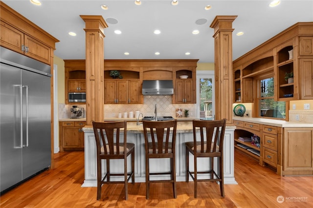 kitchen featuring ornate columns, appliances with stainless steel finishes, a breakfast bar, an island with sink, and wall chimney range hood