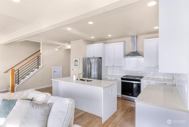 kitchen with light hardwood / wood-style flooring, wall chimney range hood, an island with sink, stainless steel appliances, and white cabinets