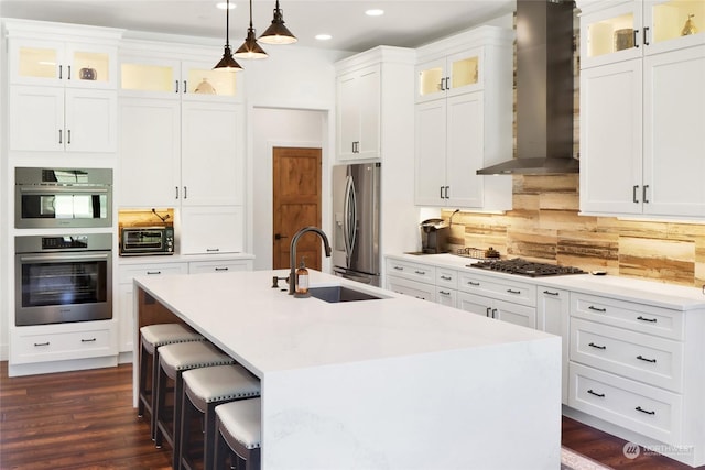 kitchen with an island with sink, sink, white cabinets, stainless steel appliances, and wall chimney exhaust hood