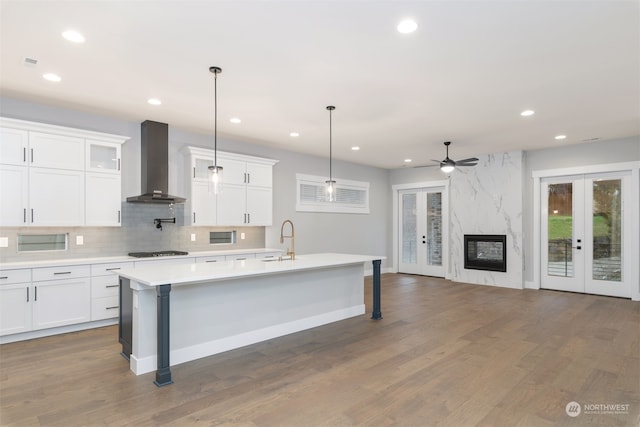 kitchen featuring decorative light fixtures, an island with sink, white cabinets, wall chimney range hood, and french doors