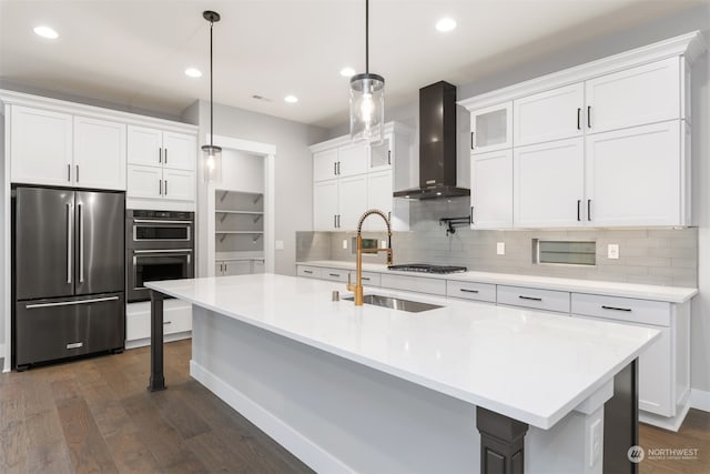 kitchen featuring wall chimney range hood, appliances with stainless steel finishes, hanging light fixtures, white cabinets, and a center island with sink