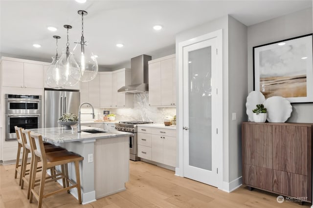 kitchen featuring an island with sink, sink, white cabinets, premium appliances, and wall chimney range hood