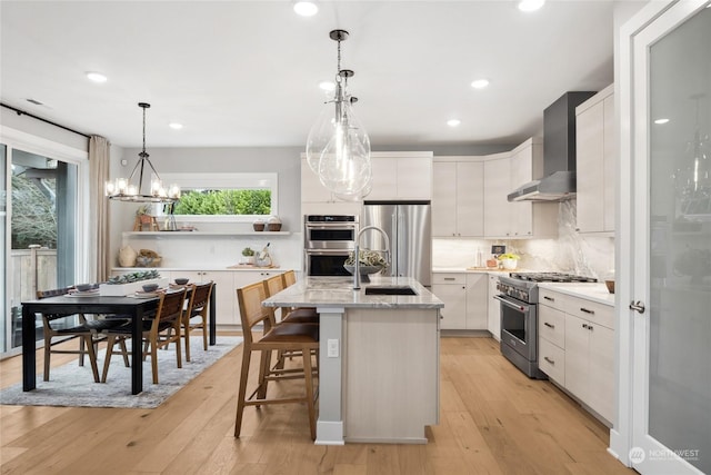 kitchen featuring appliances with stainless steel finishes, white cabinetry, an island with sink, decorative light fixtures, and wall chimney exhaust hood