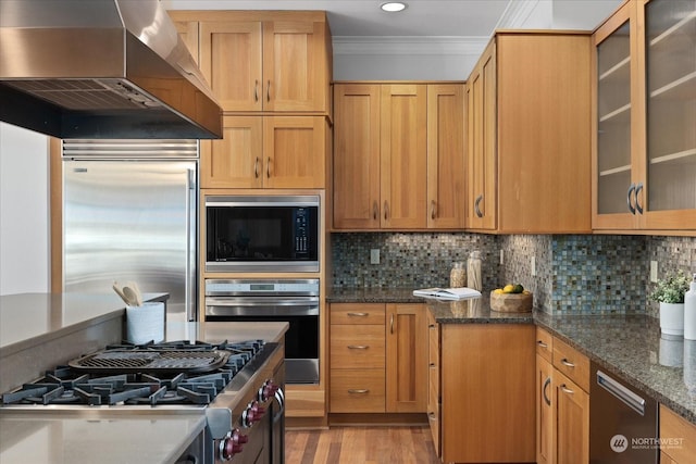 kitchen with wall chimney range hood, dark stone countertops, built in appliances, tasteful backsplash, and ornamental molding