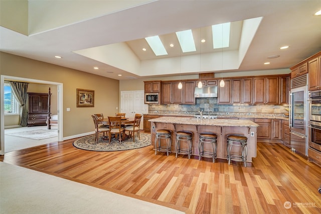 kitchen featuring under cabinet range hood, a kitchen breakfast bar, hanging light fixtures, brown cabinetry, and a center island with sink