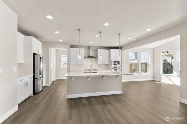 kitchen with appliances with stainless steel finishes, white cabinetry, hanging light fixtures, a kitchen island with sink, and wall chimney exhaust hood