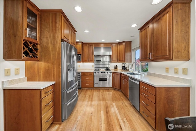 kitchen featuring a sink, light wood-style floors, appliances with stainless steel finishes, wall chimney exhaust hood, and brown cabinetry