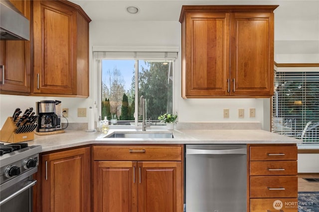 kitchen featuring brown cabinetry, a sink, stainless steel appliances, light countertops, and wall chimney exhaust hood