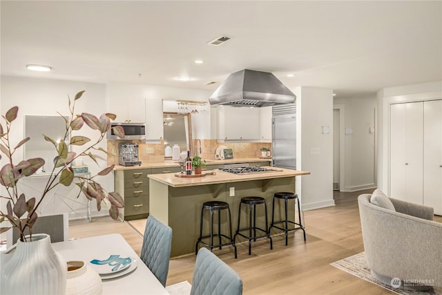 kitchen featuring white cabinetry, island range hood, light wood-type flooring, appliances with stainless steel finishes, and backsplash